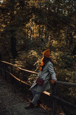 Genç bir kız sonbahar mevsiminde ahşap parkta tek başına yürüyor. Dışarıda turuncu şapkalı ve gri yün ceketli bir kadın var..