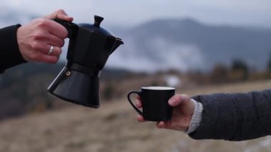 A person pouring out coffee from Moka pot into a cup outdoors in front of foggy mountains background