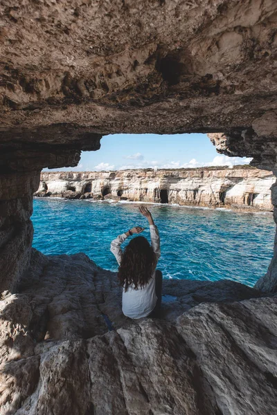 Young woman from behind is sitting in cave hole and looking at sea caves near cape Greco in national park with turquoise water sea in Ayia Napa, Cyprus.