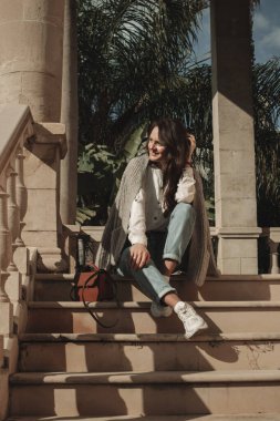 Kıbrıs Rum Kesimi 'nin Larnaka kentindeki sütunları ve tropikal bitkileri olan eski binanın yakınında oturan genç bir kadın eğleniyor..