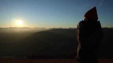Side view of a young woman in a hat drinking tea from a cup outdoors in front of mountains. Cloudy hill background in the autumn season