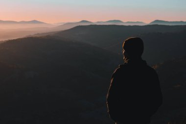 A young man from behind is standing and looking away at the mountains' autumn landscape in the morning