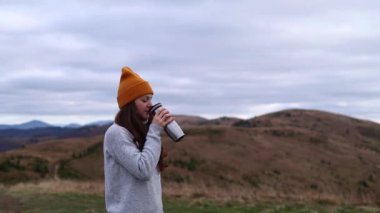 A young woman in an orange beanie hat is drinking tea from a thermos outdoors in front of mountains. Cloudy hill background in the autumn season