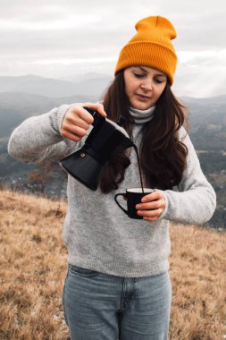 A woman in an orange beanie hat pouring out coffee from Moka pot into a cup outdoors in autumn time. Girl in a cozy casual sweater in front of a beautiful mountains view