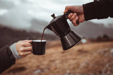 A person pouring out coffee from Moka pot into a cup outdoors in front of foggy mountains background