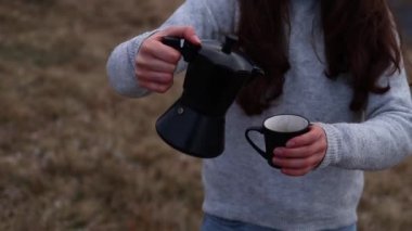 Close up of a woman pouring out coffee from Moka pot into a cup outdoors in autumn time. Girl in cozy casual sweater