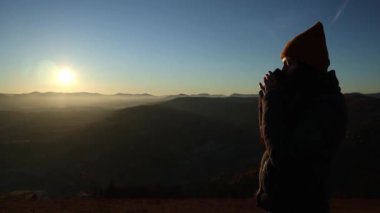 Side view of a young woman in a hat drinking tea from a cup outdoors in front of mountains. Cloudy hill background in the autumn season