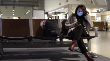 Tıbbi maskeli genç bir kadın havaalanı lobisinde oturuyor ve telefonuna bakıyor.