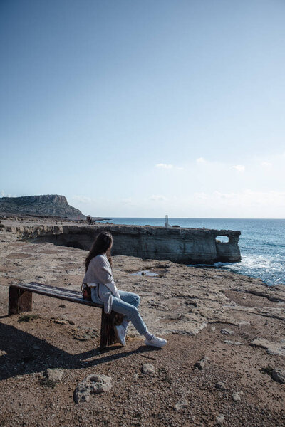 A young woman is sitting on a bench and looking at sea caves near cape Greco in a national park in Ayia Napa, Cyprus. 