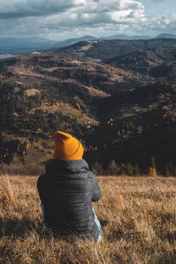 Arkadan genç bir kadın çimenlerde oturuyor ve gündüzleri dağların vadilerine bakıyor.