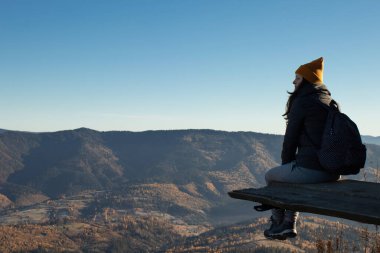 A young woman from behind with a backpack is sitting above a mountains landscape and looking away