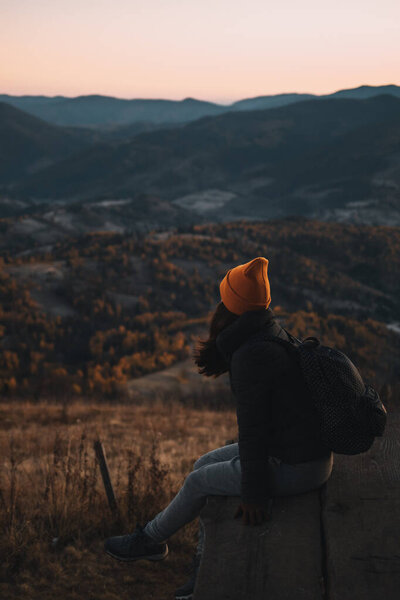 A young woman from behind with a backpack is sitting above a mountains landscape and looking away