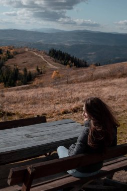 A young woman from behind is sitting on the wooden bench and looking at the mountains