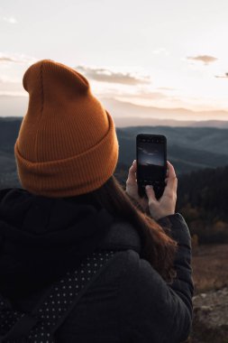 A young woman in an orange beanie is taking picture of a sunset mountains view