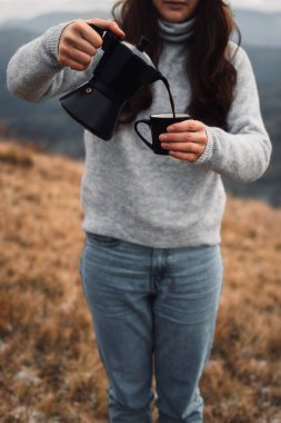 Sonbahar zamanı Moka çaydanlığından açık havada fincana kahve döken bir kadının yakın çekimi. Dağların önünde rahat bir süveter giyen kız.