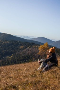 Termoslu ve sırt çantalı genç bir kadın yerde oturuyor ve dağların manzarasına bakıyor. 