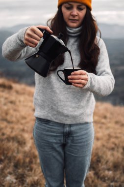 A woman in an orange beanie hat pouring out coffee from Moka pot into a cup outdoors in autumn time. Girl in a cozy casual sweater in front of a beautiful mountains view