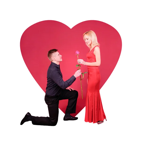Een man met roze bloem buig de knie een vrouw tegen rood hart — Stockfoto