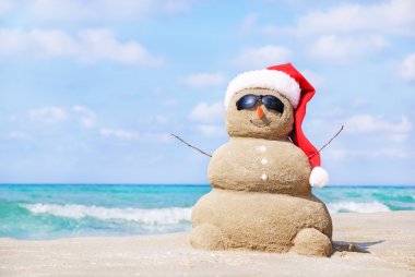 Smiling sandy snowman in red santa hat on the sea beach.