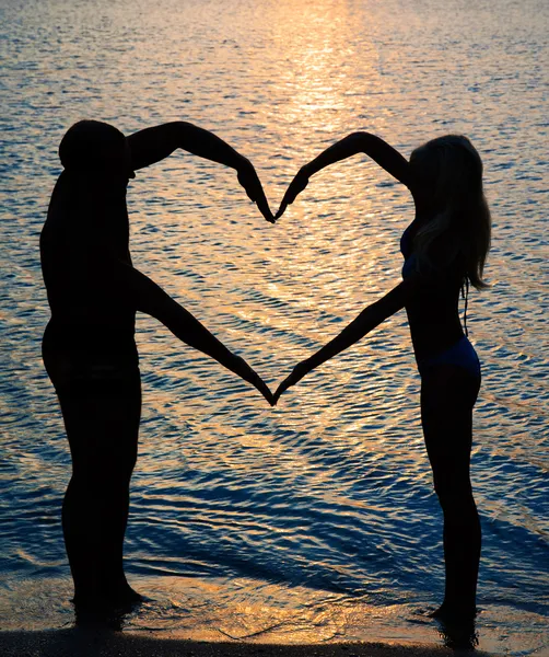 Jovem casal fazendo forma de coração com os braços na praia contra su — Fotografia de Stock