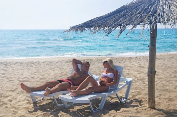 Young couple sunbathe in longue on sea beach under sunshade — Stock Photo, Image