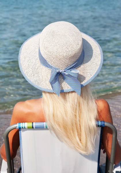 Blondýnka v bílý slaměný klobouk sedí na pláži židli a vzhled — Stock fotografie