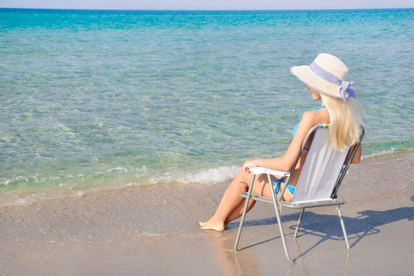 Mujer rubia en sombrero de paja blanca sentada en la silla de playa y mira —  Fotos de Stock