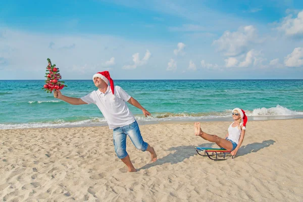 Jeune couple courant à la plage de la mer dans chapeaux Santa avec traîneau et ch — Photo