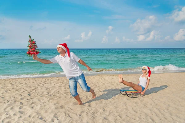 Young couple running at sea beach in santa hats with sled and ch — Stock Photo, Image