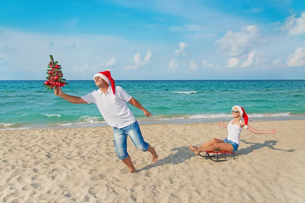 Young couple running at sea beach in santa hats with sled and ch — Stock Photo, Image