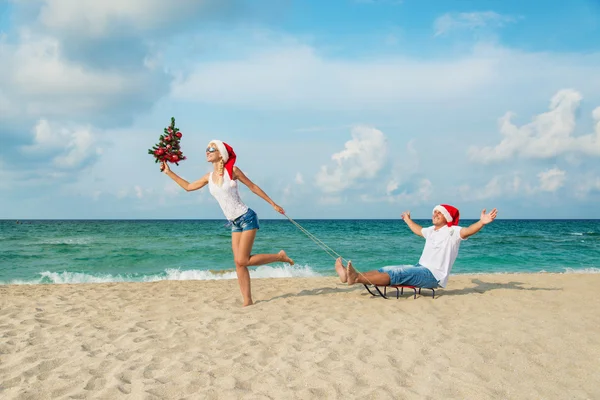 Young couple running at sea beach in santa hats with sled and ch — Stock Photo, Image