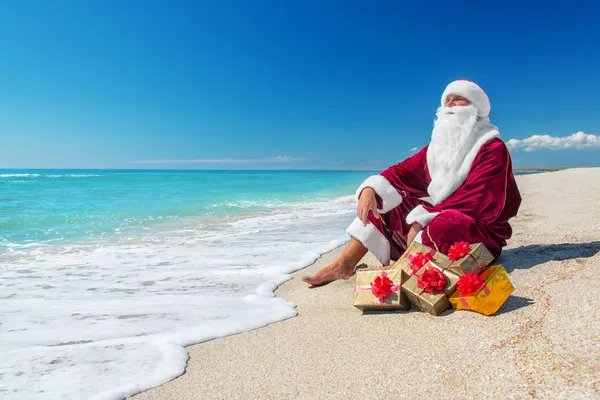 Père Noël avec de nombreux cadeaux dorés relaxant à la plage — Photo