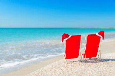 Santa hat on chaise longues at white sand beach against the sea clipart