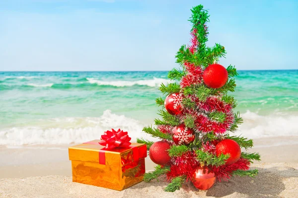 Weihnachtsbaum und goldenes Geschenk mit großer roter Schleife am Meeresstrand — Stockfoto