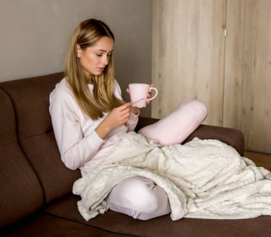 Güzel hasta bir kız elinde bir termometre ve bir fincan çay tutuyor. Ev kıyafetleri içindeki mutsuz kadın hasta, tedavi ediliyor ve ilaç içiyor. Ateşi, burnu ve ilacı akan hasta bir kadın. Evde tabletlerle tedavi