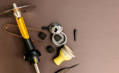 Siyah ve sarı bir nargile kahverengi bir masanın üzerinde sigara içilen aksesuarlar ve kömürle birlikte yatar. Hindistan cevizi kömürü, sarı kase, nargile salonda maşa.