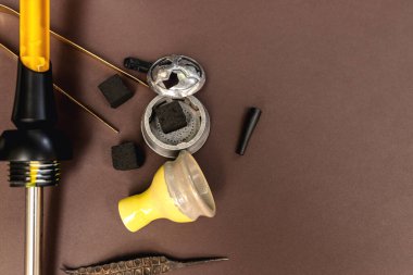 Siyah ve sarı bir nargile kahverengi bir masanın üzerinde sigara içilen aksesuarlar ve kömürle birlikte yatar. Hindistan cevizi kömürü, sarı kase, nargile salonda maşa.