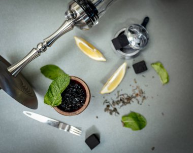 Limonlu aromatik tütün ve gri masada sigara içilen aksesuarlı nane şekeri kasesi. Nargile, nargile tüttürüyor. Çelik nargile, maşa, çay ve hindistan cevizi kömürü.