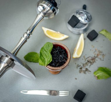 Limonlu aromatik tütün ve gri masada sigara içilen aksesuarlı nane şekeri kasesi. Nargile, nargile tüttürüyor. Çelik nargile, maşa, çay ve hindistan cevizi kömürü.