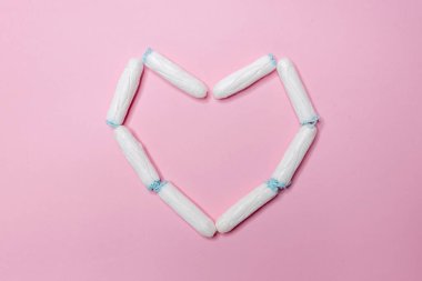 Birçok beyaz hijyenik tampon açık pembe arka planda kalp şeklinde yatar. Adet döngüsü ve hamilelik. Negatif hamilelik testi ve doğum kontrol hapları.