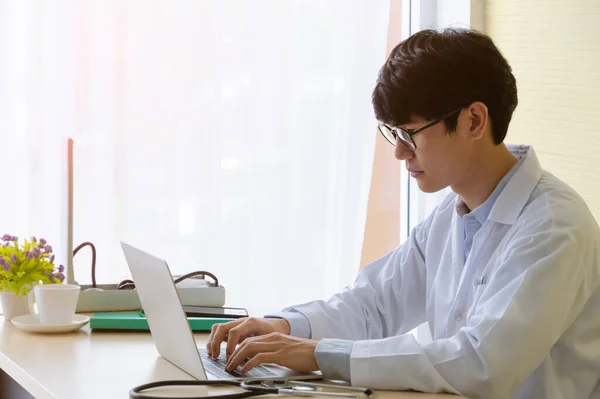 Young doctor physician wearing white uniform and using laptop in hospital office. Male doctor in eyeglasses typing on his computer at desk