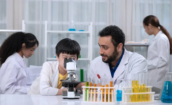 Kid boy wearing white laboratory coat and learning how to use microscope in science laboratory room with scientist teacher. Children education concept with experiment, fun and enjoy class.