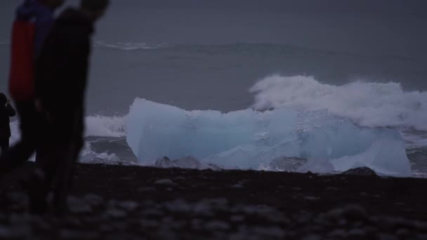 波はダイヤモンドビーチで氷山の上に砕け、観光客はゆっくりと交差します。 — ストック動画