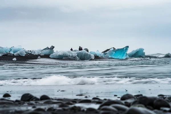 Blaue Eisberge über dem schwarzen Sandstrand mit verschwommenen Touristen — Stockfoto