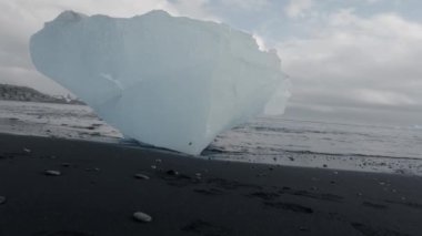 Ağır çekimde siyah kumun üzerinde kocaman mavi bir buzdağı.