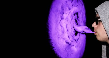 Kapüşonlu ve güneş gözlüklü bir adam e-sigara içiyor ve siyah arka planda mor bir bulut üflüyor..