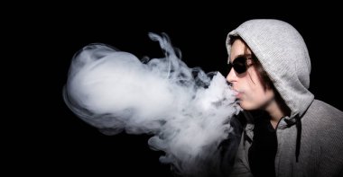 Kapüşonlu ve güneş gözlüklü bir adam e-sigara içiyor ve siyah arka planda bir bulut üflüyor.