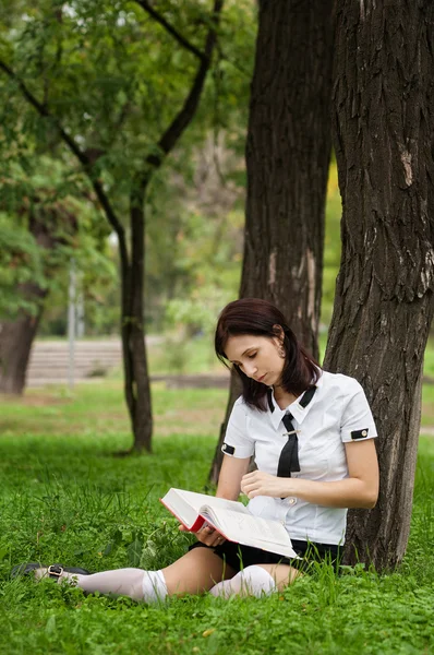 Estudiante leyendo el libro. Hermosa joven con libro sentado en la hierba y apoyado en el árbol. Exterior . — Foto de Stock