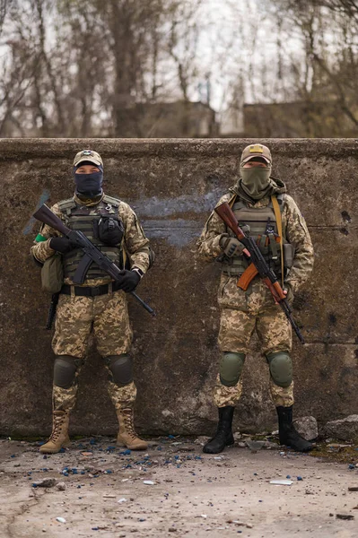 Two Ukrainian soldiers in military uniforms and with machine guns in front of a concrete wall