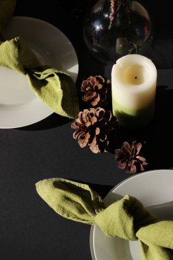 Noel Yemeği Servisi. Yeşil Peçeteli Aydınlık Tabaklar Kara Arkaplanda Mumlar ve Koniler ile. Yeni yıl, Noel arifesi Kutlaması.